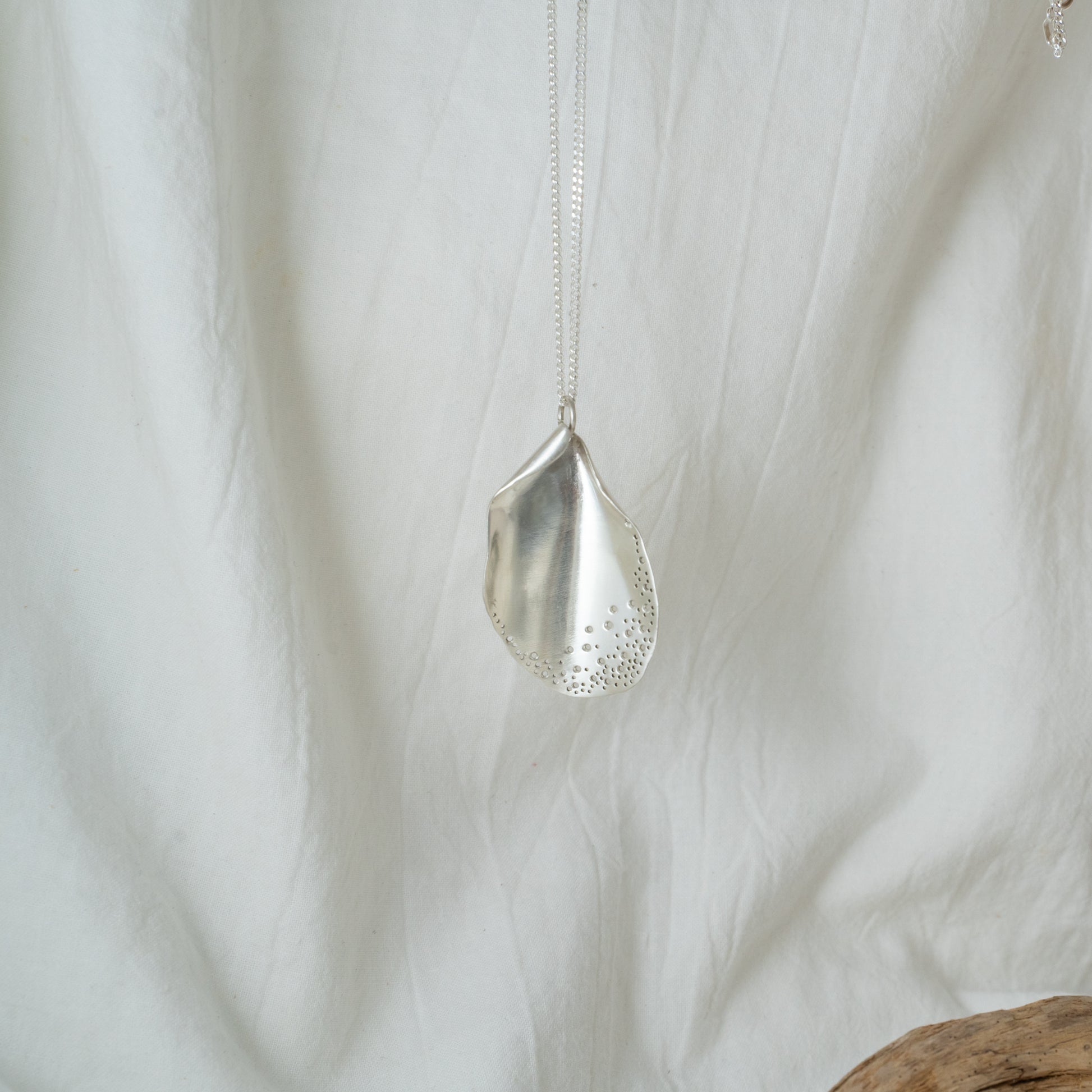 A silver pendant necklace with a chain, featuring hand-drilled holes and a solid silver curb chain, displayed against a white fabric backdrop.