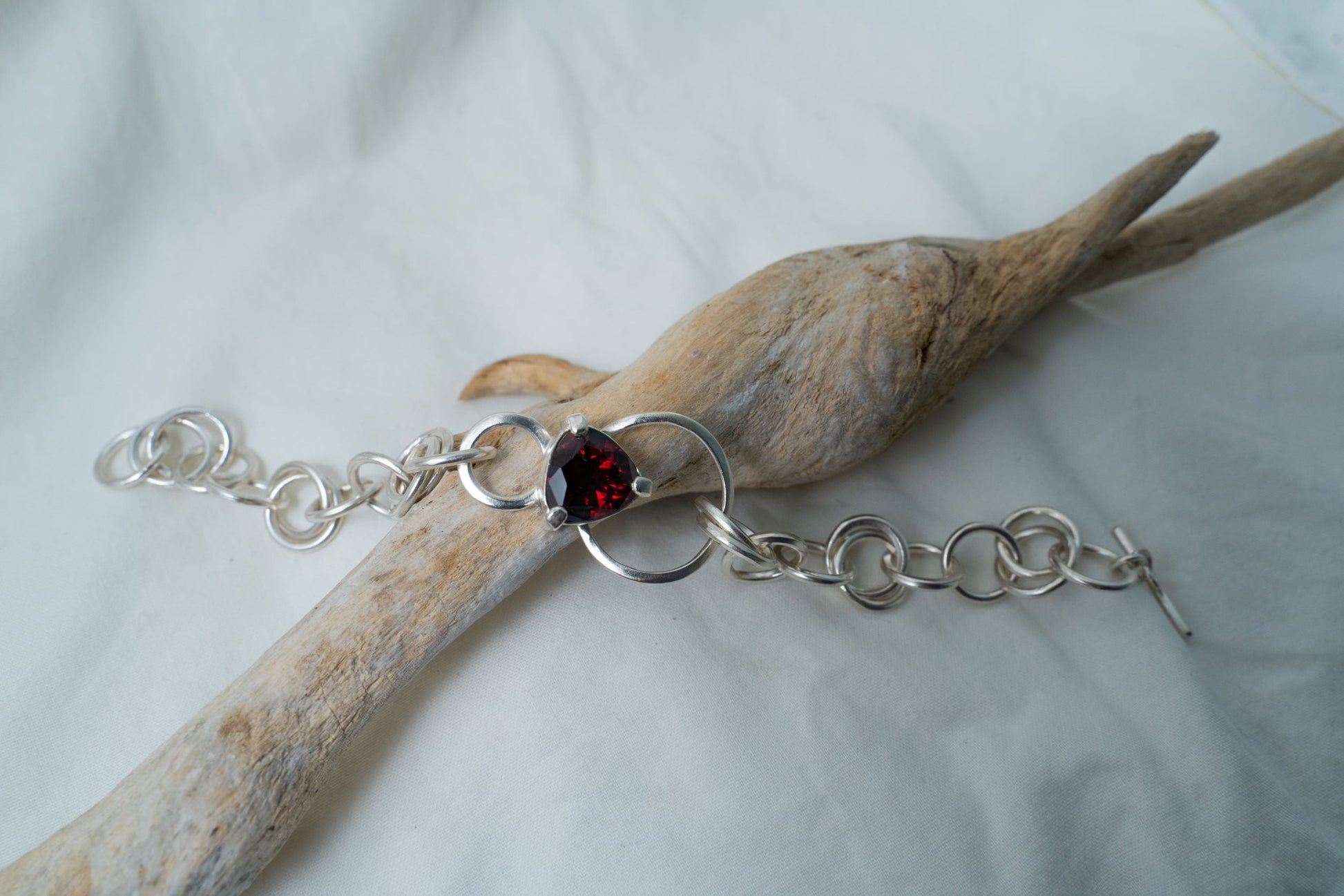 A handcrafted silver chain bracelet is photographed placed on a piece of driftwood found in Hong Kong. The camera is aimed at the trilliant cut garnet showing off the flashes of deep red within the gemstone