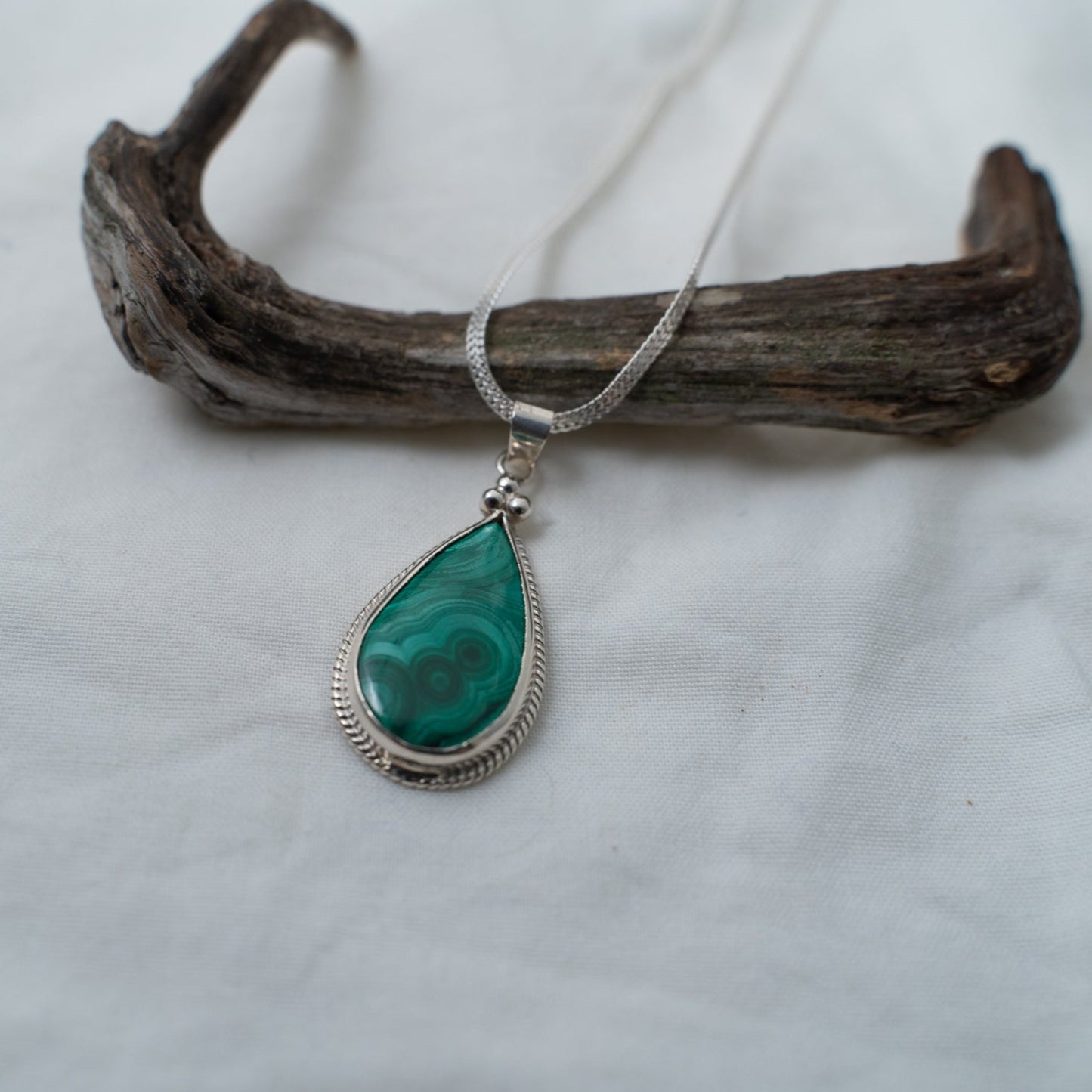A photograph of a beautiful, deep green, natural malachite gemstone that is set in a silver, wire bezel. This pendant is in the shape of a teardrop and has three, triangular, silver, granules sitting at the top of the teardrop. It shows where the chain goes and you can see a standard silver bail at the top of the pendant for the chain. The pendant is photographed on a white background, whilst propped against a piece of driftwood.
