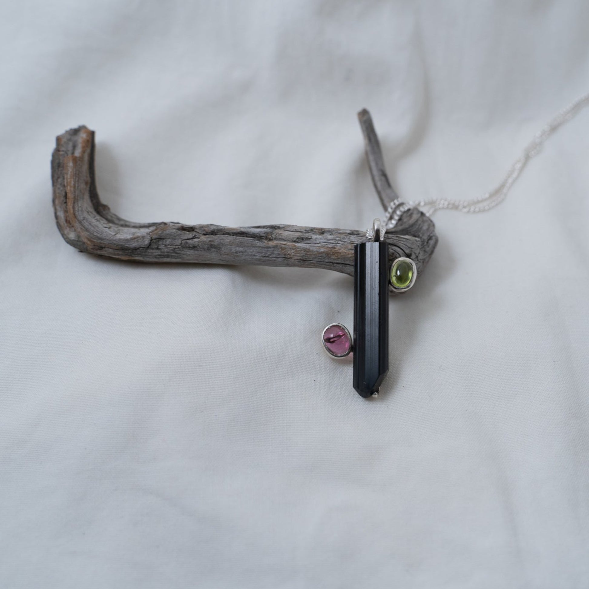 The black and pink tourmaline with a green peridot pendant for sale is propped against a piece of driftwood and photographed against a white back drop. This shows you what the pendant looks like in depth from a direct head on approach.