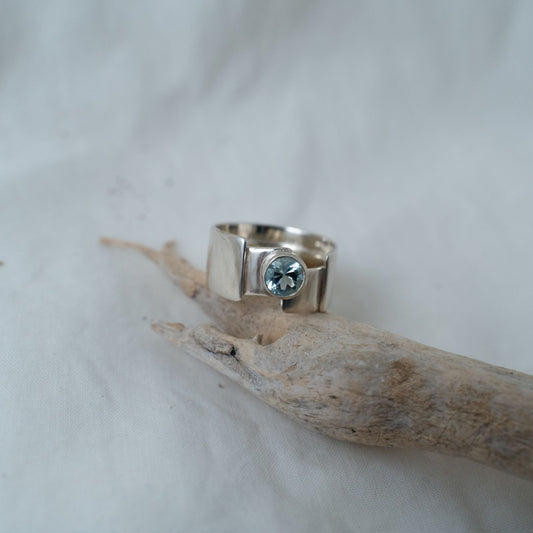 A photograph of the gender-fluid, chunky, round cut aquamarine, silver ring. This is a larger ring in both width and ring size. It is set with a bezel setting and wraps around the finger with a large thick, wide ring shank. 