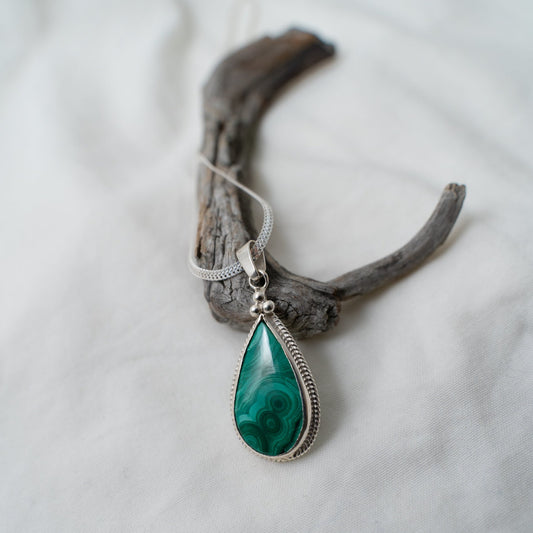 A photograph of a beautiful, deep green, natural malachite gemstone that is set in a silver, wire bezel. This pendant is in the shape of a teardrop and has three, triangular, silver, granules sitting at the top of the teardrop. It shows where the chain goes and you can see a standard silver bail at the top of the pendant for the chain. The pendant is photographed on a white background, whilst propped against a piece of driftwood. 